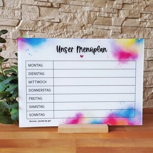 Könnte beinhalten: Ein weißes Trockenlöschboard mit einem bunten Aquarellhintergrund. Das Board hat einen schwarzen Rahmen und einen schwarzen Titel, der "Unser Menüplan" mit einem kleinen rosa Herz sagt. Das Board ist in sieben Abschnitte unterteilt, die jeweils mit einem Wochentag in Deutsch beschriftet sind: Montag, Dienstag, Mittwoch, Donnerstag, Freitag, Samstag, Sonntag. Das Board steht auf einem Holzständer.