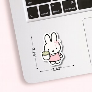 May include: A sticker featuring a cartoon bunny holding a cup of coffee. The bunny is white with a pink bow and dress. The sticker measures 2.16 inches by 1.43 inches.