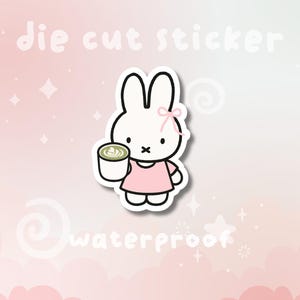 May include: Die cut sticker featuring a white bunny wearing a pink dress and holding a cup of matcha. The sticker has a pink bow and the word "waterproof" is written on the bottom. The background is pink.