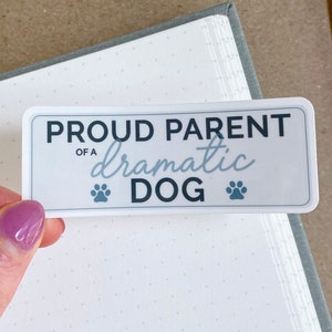 May include: White sticker with blue text that reads "Proud Parent of a dramatic DOG" with two paw prints on either side of the word "DOG".