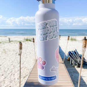 Sticker on white water bottle at the beach with other dog-themed decals in the background.