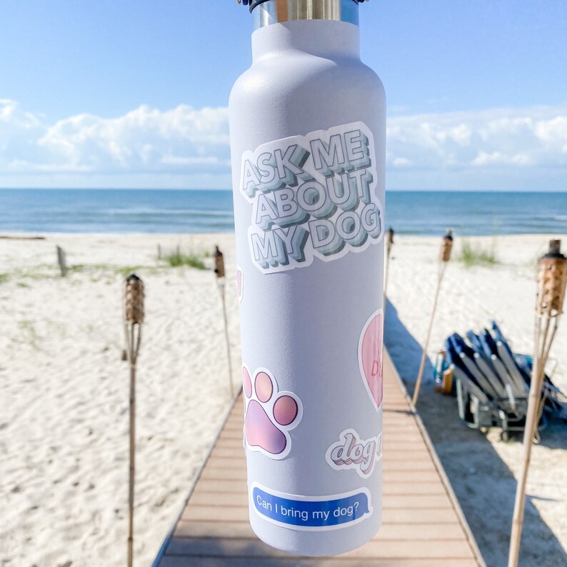 Sticker on white water bottle at the beach with other dog-themed decals in the background.