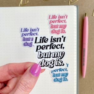 May include: A white sticker with black text that reads "Life isn't perfect, but my dog is." The sticker is being held by a hand with pink nail polish.