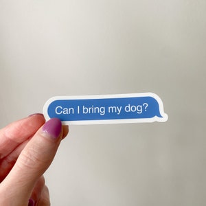 Hand holding “Can I bring my dog?” sticker in front of soft neutral backdrop.