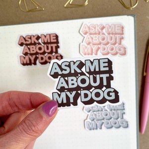 A hand with purple nail polish holds a sticker that says "Ask Me About My Dog." Other similar stickers are on a dotted notebook and a pink pen is nearby.