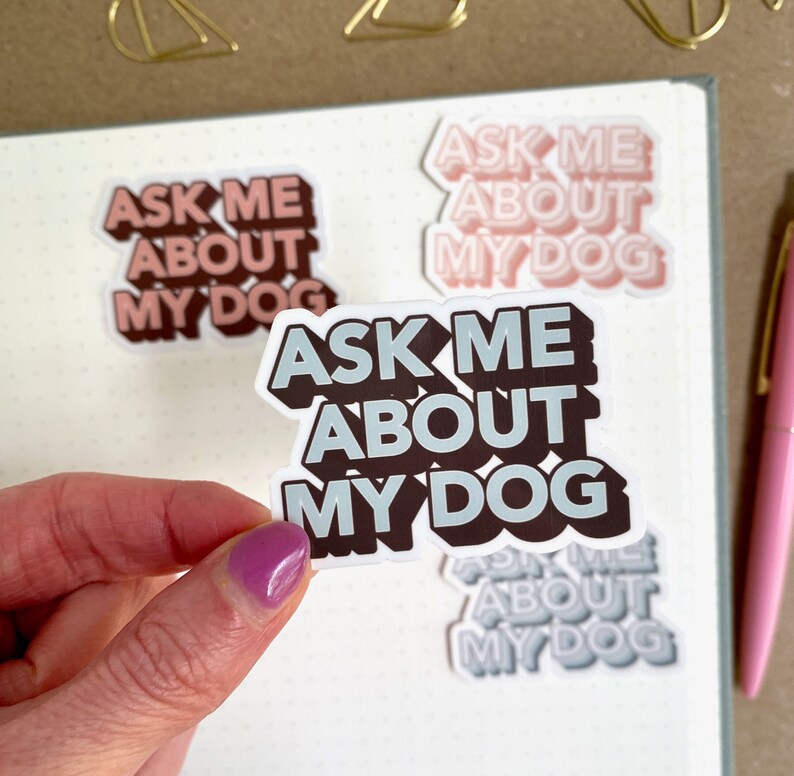 A hand with purple nail polish holds a sticker that says "Ask Me About My Dog." Other similar stickers are on a dotted notebook and a pink pen is nearby.