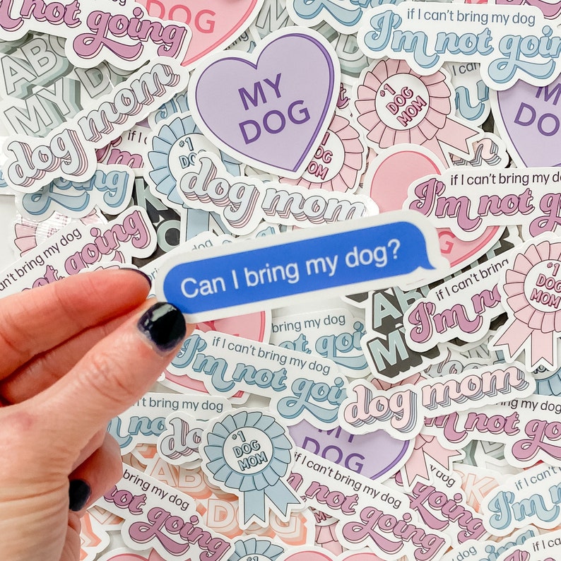 Hand holding sticker in front of colorful pile of dog mom-themed stickers including “If I can't bring my dog, I’m not going,” “I love MY DOG,” and “dog mom.”
