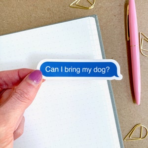 Hand holding “Can I bring my dog?” sticker on white dotted journal page with gold paperclips and pink pen.