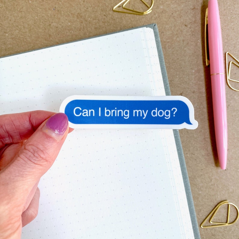 Hand holding “Can I bring my dog?” sticker on white dotted journal page with gold paperclips and pink pen.