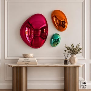 Puede incluir: Tres esculturas de pared redondeadas y reflectantes en magenta, naranja y turquesa. Debajo, una mesa consola sostiene una pila de libros con las etiquetas MIAMI, NEW YORK y CAPRI, un cuenco, un difusor de varillas y un jarrón con follaje.