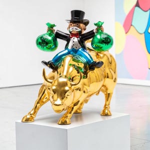 May include: A glossy gold bull sculpture with a figure riding on its back. The figure wears a black top hat, a white shirt with a red bowtie, and a black jacket, holding two bright green money bags with black dollar signs printed on them.