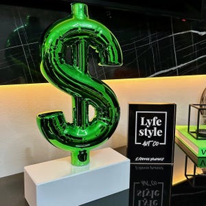 May include: A green, shiny, inflated dollar sign sculpture on a white base. The sculpture is on a table with a black and white sign that reads "Lyfe Style Art Co. Express Yourself. Express Yourself."