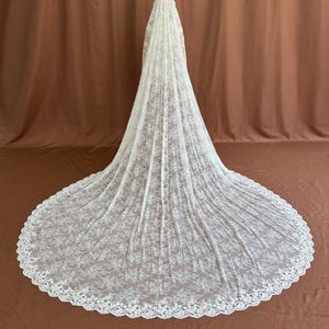 May include: A long, ivory-colored wedding veil with a delicate floral lace pattern. The veil cascades down to the floor, creating a dramatic train. The edges are scalloped, adding a refined touch. The background is a solid brown.