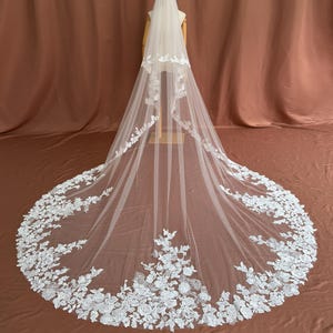 Puede incluir: Un velo de novia blanco y largo con intrincados detalles de encaje floral en los bordes. El velo se exhibe en un maniquí, mostrando su diseño fluido y su delicada artesanía. La cola del velo se extiende con gracia.