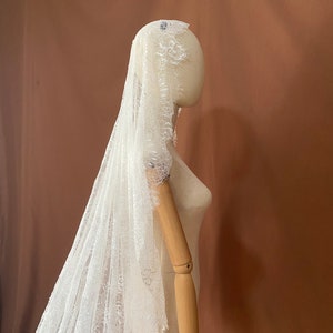 May include: A white lace bridal veil, draped over a mannequin. The veil features intricate floral lace detailing along the edges and is displayed against a brown backdrop. The delicate fabric creates a soft, romantic look, perfect for weddings.