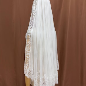 Puede incluir: Un velo de novia blanco con intrincados detalles de encaje. El velo es largo y fluido, cayendo por la espalda. El encaje es visible en los bordes y las mangas, agregando un toque de elegancia. El velo se muestra en un maniqu&iacute;.