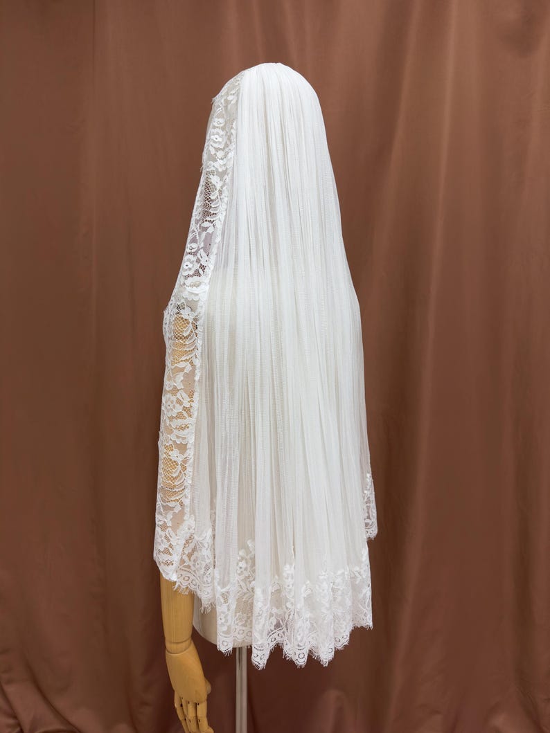 Puede incluir: Un velo de novia blanco con intrincados detalles de encaje. El velo es largo y fluido, cayendo por la espalda. El encaje es visible en los bordes y las mangas, agregando un toque de elegancia. El velo se muestra en un maniqu&iacute;.