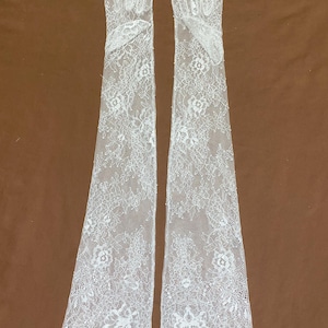 May include: A pair of elegant, white lace gloves. The gloves are long, extending well past the wrist, and feature a delicate floral pattern. The lace is intricately detailed, with scalloped edges. The gloves are displayed against a brown background.