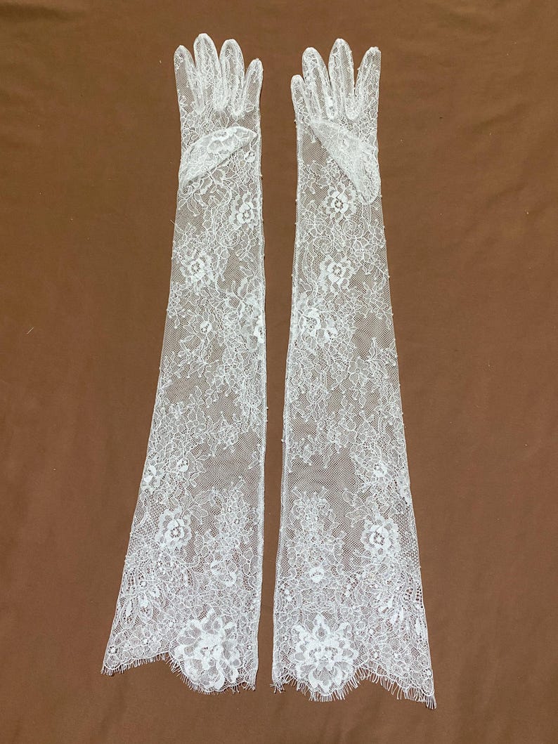 May include: A pair of elegant, white lace gloves. The gloves are long, extending well past the wrist, and feature a delicate floral pattern. The lace is intricately detailed, with scalloped edges. The gloves are displayed against a brown background.