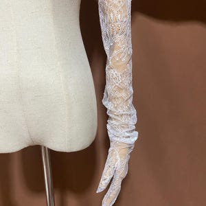 Puede incluir: Guantes largos de encaje blanco con un delicado estampado floral. Los guantes se extienden por el brazo y presentan un diseño sin dedos. El encaje está intrincadamente detallado, creando un aspecto romántico y elegante. Los guantes se exhiben en un brazo de maniquí.