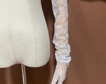 May include: White lace gloves with a delicate floral pattern. The gloves are long, extending up the arm, and feature a fingerless design. The lace is intricately detailed, creating a romantic and elegant look. The gloves are displayed on a mannequin arm.