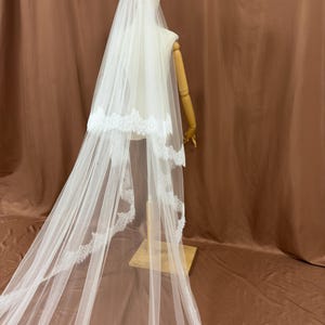 Puede incluir: Un velo de novia blanco con un delicado ribete de encaje. El velo est&aacute; drapeado sobre un maniqu&iacute;, mostrando su longitud y dise&ntilde;o. El velo est&aacute; hecho de una tela transparente, creando un aspecto suave y elegante para una boda.