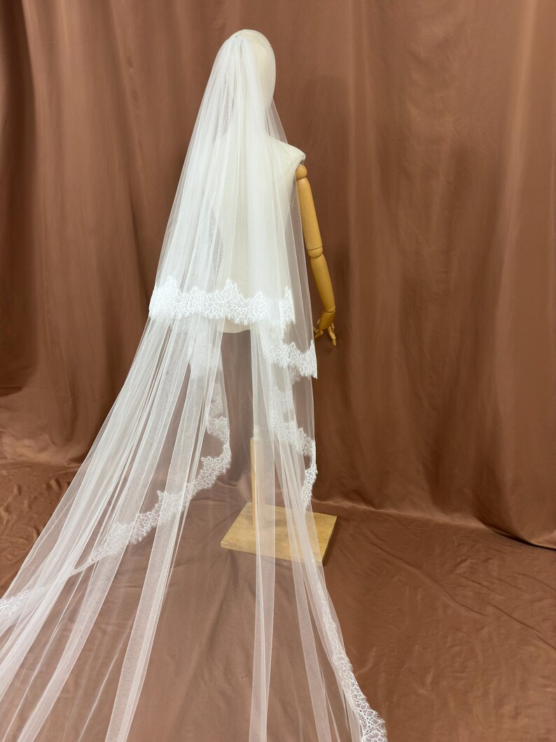 Puede incluir: Un velo de novia blanco con un delicado ribete de encaje. El velo est&aacute; drapeado sobre un maniqu&iacute;, mostrando su longitud y dise&ntilde;o. El velo est&aacute; hecho de una tela transparente, creando un aspecto suave y elegante para una boda.