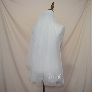 May include: A white tulle bridal veil with embroidered initials "W & T" at the hem. The veil is draped over a mannequin, showcasing its delicate, layered design. The background is a neutral beige.