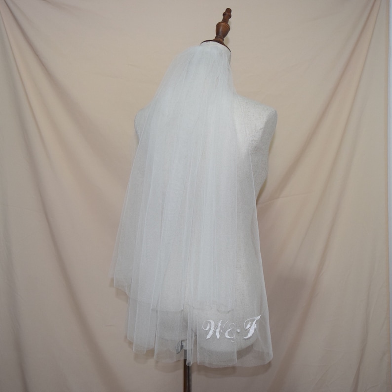 May include: A white tulle bridal veil with embroidered initials "W & T" at the hem. The veil is draped over a mannequin, showcasing its delicate, layered design. The background is a neutral beige.