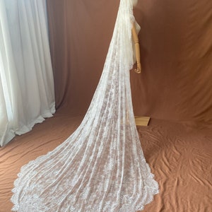 May include: A long, white lace wedding veil. The veil is displayed on a mannequin, showcasing its intricate floral lace pattern and scalloped edge. The veil cascades down to the floor, creating a dramatic effect.