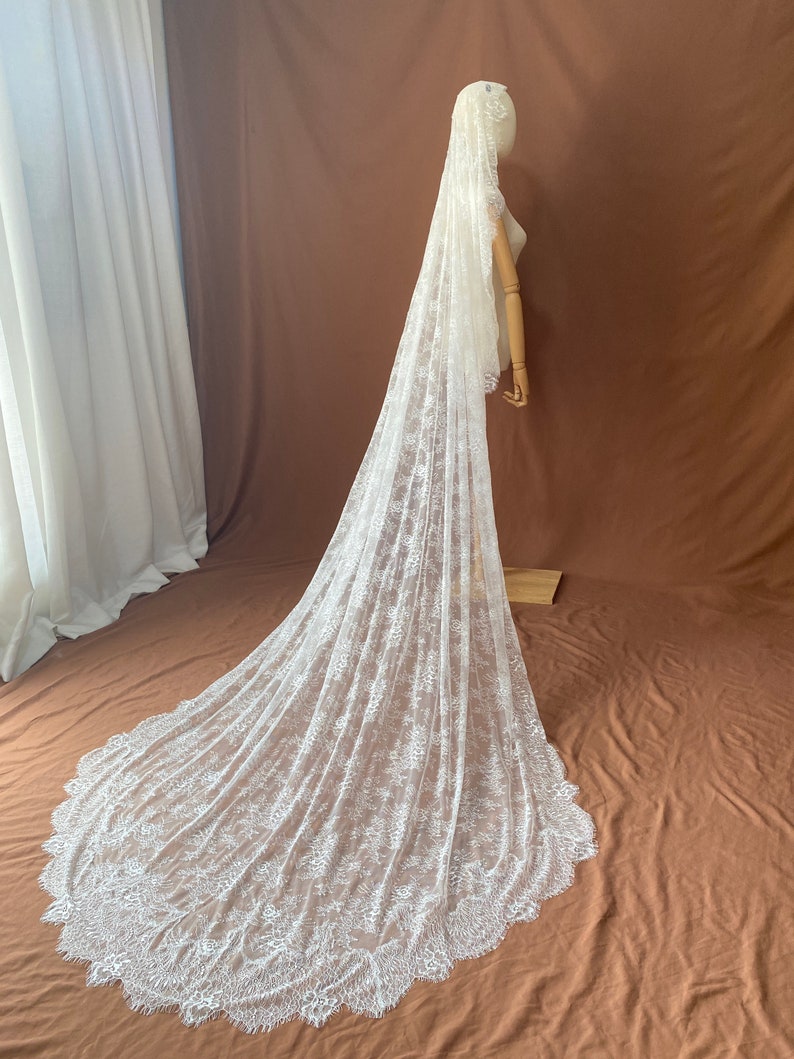 May include: A long, white lace wedding veil. The veil is displayed on a mannequin, showcasing its intricate floral lace pattern and scalloped edge. The veil cascades down to the floor, creating a dramatic effect.