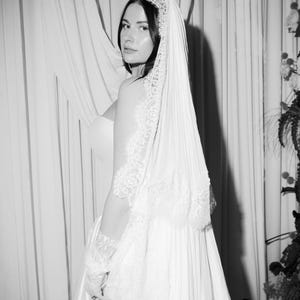 Puede incluir: Fotograf&iacute;a en blanco y negro de una novia con un vestido de novia blanco y un velo. El velo presenta delicados detalles de encaje, y el vestido tiene un dise&ntilde;o simple y elegante. La novia mira por encima del hombro.