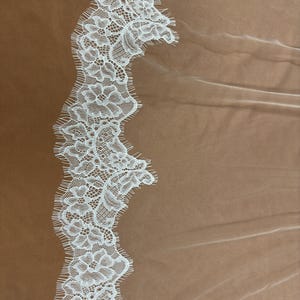 Puede incluir: Primer plano de un ribete de encaje blanco con un dise&ntilde;o floral. El encaje presenta patrones intrincados y un borde festoneado. El fondo es una tela transparente de color marr&oacute;n claro.