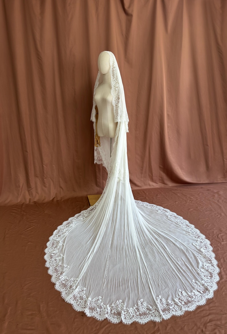 Puede incluir: Un velo de novia blanco con una larga cola, exhibido en un maniqu&iacute;. El velo presenta un delicado ribete de encaje y un dise&ntilde;o plisado y fluido. El maniqu&iacute; se encuentra sobre un fondo marr&oacute;n.