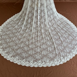 May include: A white lace wedding veil with a floral pattern and a scalloped edge. The veil is spread out on a brown surface, showcasing its full, circular shape. The delicate fabric creates a soft, romantic look.