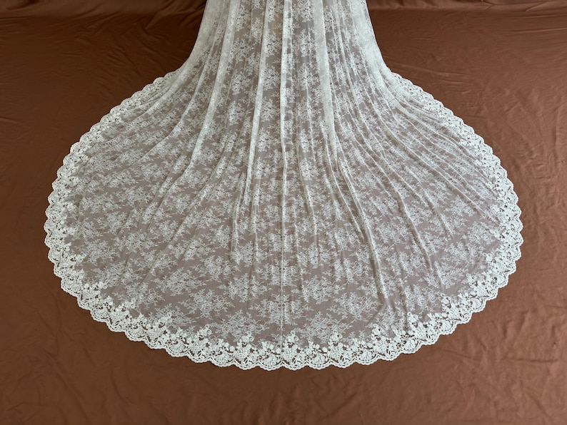 May include: A white lace wedding veil with a floral pattern and a scalloped edge. The veil is spread out on a brown surface, showcasing its full, circular shape. The delicate fabric creates a soft, romantic look.
