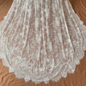 May include: White lace fabric with a floral pattern and scalloped edge. The delicate lace is semi-sheer, with a detailed design of flowers and leaves. The fabric is likely used for bridal wear or other formal garments.