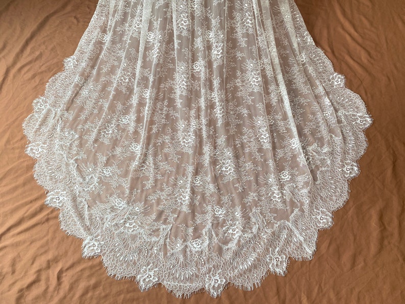 May include: White lace fabric with a floral pattern and scalloped edge. The delicate lace is semi-sheer, with a detailed design of flowers and leaves. The fabric is likely used for bridal wear or other formal garments.