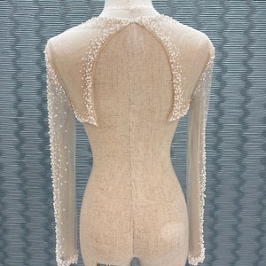 May include: A sheer, long-sleeved garment with a high neckline and a decorative pattern of small, white beads. The back features a keyhole design, and the garment is displayed on a mannequin.