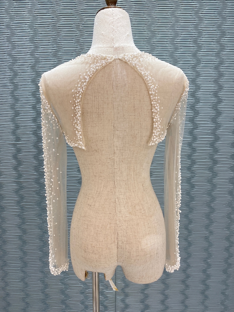May include: A sheer, long-sleeved garment with a high neckline and a decorative pattern of small, white beads. The back features a keyhole design, and the garment is displayed on a mannequin.