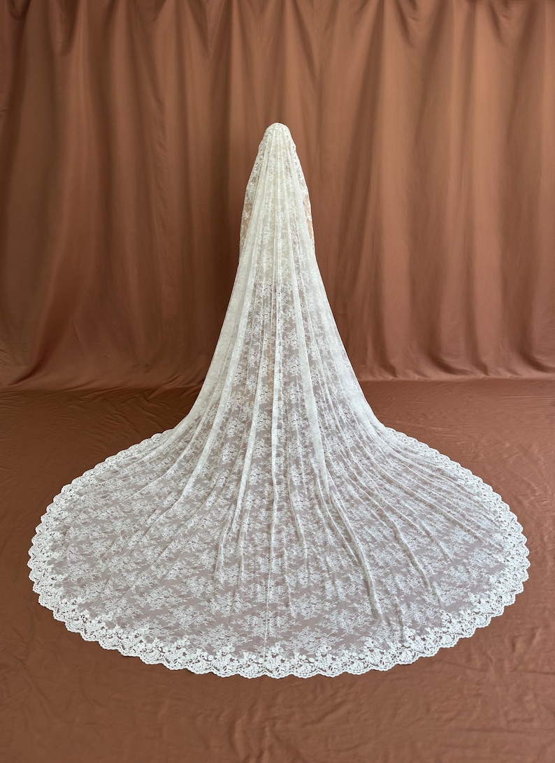 May include: A long, ivory-colored wedding veil with a delicate floral lace pattern. The veil cascades down to the floor, creating a dramatic train. The edges are scalloped, adding a refined touch. The background is a solid brown.