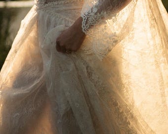 Puede incluir: Un vestido de encaje color marfil de manga larga con intrincados bordados florales, un corpiño ajustado con escote en V y una falda fluida. El delicado tejido de encaje tiene una calidad semitransparente con bordes festoneados en las mangas.