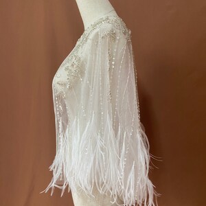 Puede incluir: Una capa blanca y transparente adornada con cuentas y plumas, exhibida en un maniqu&iacute;. El escote y los hombros presentan un intrincado trabajo de cuentas. Hileras de cuentas cuelgan de la capa, y las mangas est&aacute;n adornadas con plumas blancas.