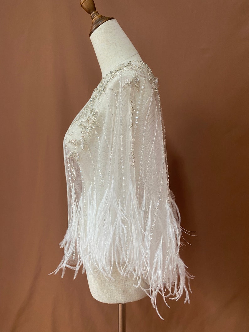 Puede incluir: Una capa blanca y transparente adornada con cuentas y plumas, exhibida en un maniqu&iacute;. El escote y los hombros presentan un intrincado trabajo de cuentas. Hileras de cuentas cuelgan de la capa, y las mangas est&aacute;n adornadas con plumas blancas.