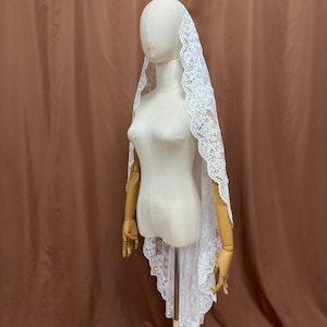 May include: A white lace veil with a scalloped edge is draped over a mannequin. The veil is made of delicate lace and has a floral pattern. The mannequin is on a wooden stand, and the background is a solid brown color.