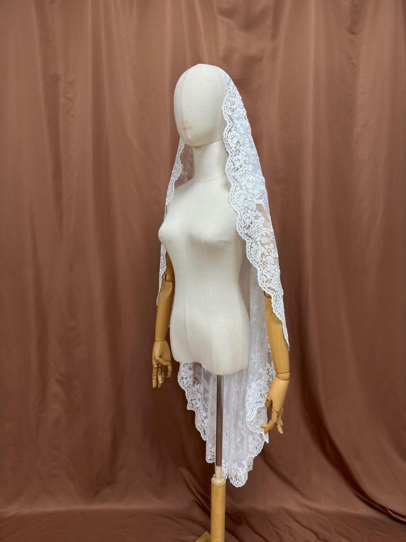 May include: A white lace veil with a scalloped edge is draped over a mannequin. The veil is made of delicate lace and has a floral pattern. The mannequin is on a wooden stand, and the background is a solid brown color.