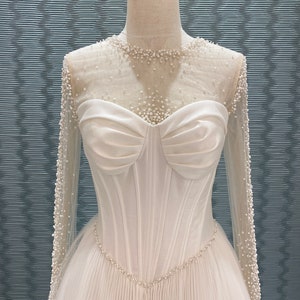May include: A white wedding dress with a sweetheart neckline and long sleeves. The bodice is fitted with vertical seams and a pearl-beaded trim. The sheer sleeves and neckline are adorned with small pearls. The skirt is pleated.