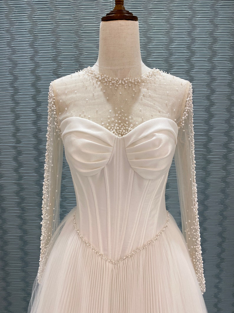 May include: A white wedding dress with a sweetheart neckline and long sleeves. The bodice is fitted with vertical seams and a pearl-beaded trim. The sheer sleeves and neckline are adorned with small pearls. The skirt is pleated.