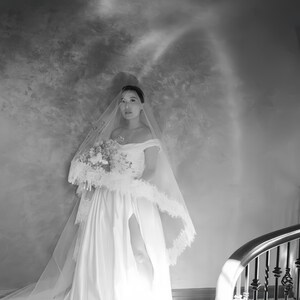 May include: A bride in a white off-the-shoulder wedding dress with a high slit, holding a bouquet. She wears a veil with lace trim. The background is a textured wall with a staircase in the corner.