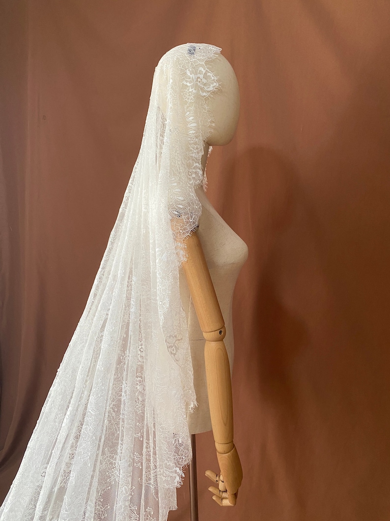 May include: A white lace bridal veil, draped over a mannequin. The veil features intricate floral lace detailing along the edges and is displayed against a brown backdrop. The delicate fabric creates a soft, romantic look, perfect for weddings.